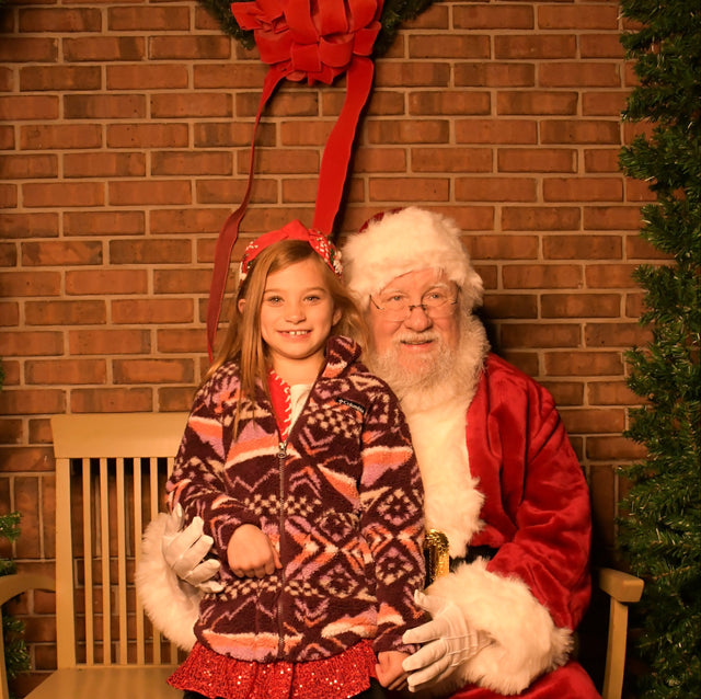 Santa Claus sitting on a bench with a young girl in front of a brick wall with a wreath.